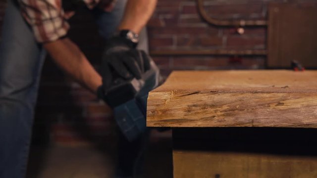 close up shot of the worker's hand, who is leveling the edges at the table in the carpentry workshop, uses an electric grinding machine for working with wood