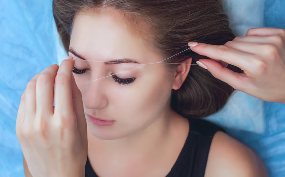 A make-up artist plucks an eyebrow with a thread from a beautiful brunette girl in a beauty salon.
