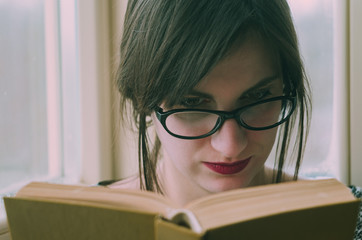 Portrait of young attractive brunette woman wearing glasses, reading book, sitting near the window
