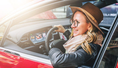 Side view, close up. Young smiling woman in glasses and hat sits behind wheel in red car. Trip, caravanning, tourism, journey.