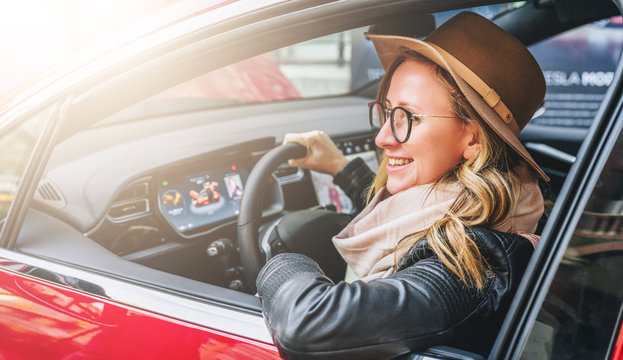 Side view, close up. Young smiling woman in glasses and hat sits behind wheel in red car. Trip, caravanning, tourism, journey.