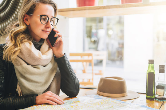 Young female tourist in eyeglasses is sitting in cafe at table and talking on cell phone. Girl is calling her friend on phone. There is map and hat on table. Tourism, travel, vacation.