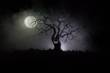 Silhouette of scary Halloween tree with horror face on dark foggy toned background with moon on back side. Scary horror tree with zombie and demon faces.