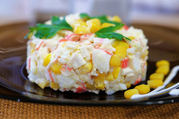 On a dark plate salad of crab sticks, corn, boiled rice, eggs and with the addition of mayonnaise. The dish is decorated with leaves of green parsley. Close up view.