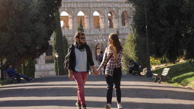 Young happy couple walk in park road with trees colosseum in background in rome at sunset lovely beautiful girl with long hair holding hands slow motion