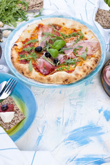 Round large pizza from the oven with prosciutto and olives, arugula and basil. Table setting for lunch or dinner. Table and dishes in blue. Free space for text.