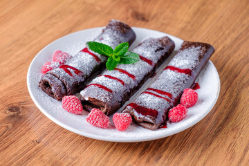 On a wooden table a plate with rolled brown pancakes with the addition of cocoa. Pancakes are poured with jam and decorated with green mint leaves and raspberries.
