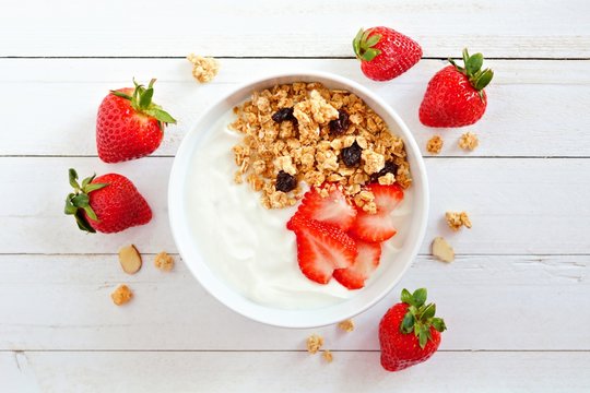 Bowl of yogurt with strawberries and granola, over a white wood background. Scattered berries.