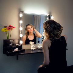 Attractive blonde girl primp at the mirror with lamps in the beauty studio. A woman in a beauty salon admires her reflection. Beautiful face with natural makeup and luxurious curls.
