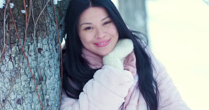 Portrait of a beautiful Asian woman against the background of the snow park. Snowflakes lie on her hair. She is dressed in a pink coat and white gloves. She holds hair and smile to the camera.