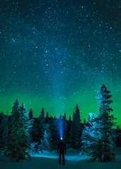 Selbstklebende Fototapeten Mann steht unter Nordlicht im Winterwald mit Scheinwerferstrahl und Sternenhimmel © Albert