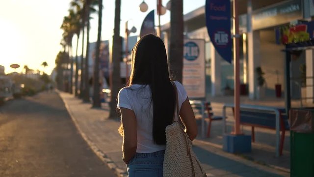 Back view of brunette young woman wearing t-shirt walking down street on sunny summer day in Cyprus. She is going to the beach to enjoy beautiful sunrise.