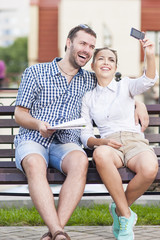 Travel Ideas and Concepts. Young Caucasian Couple in Love Sitting on Bench Outdoors While Taking Selfie Pictures.
