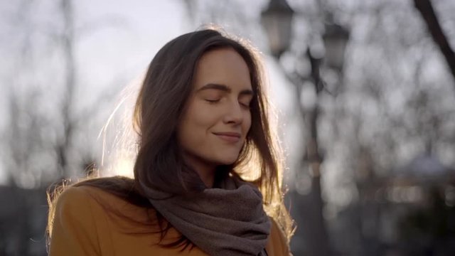 Portrait of beautiful woman with brown long hair wearing stylish coat and scarf taking deep breath, enjoying walk in city park in slow motion. Film look
