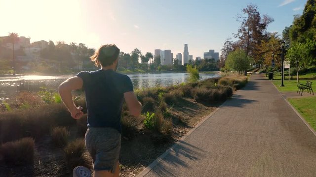 Athletic man jogs around a small lake in Los Angeles. Buildings of Los Angeles skyline can be seen in the distance.