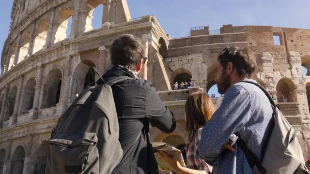 Three young friends tourists standing in front of colosseum in rome reading map guide for directions pointing with backpacks sunglasses happy beautiful girl long hair slow motion