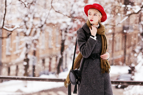 Outdoor portrait of young beautiful girl wearing long grey winter coat, red hat, olive scarf, with small round leather bag. Model walking in street of european city. Snowfall. Copy, empty space  