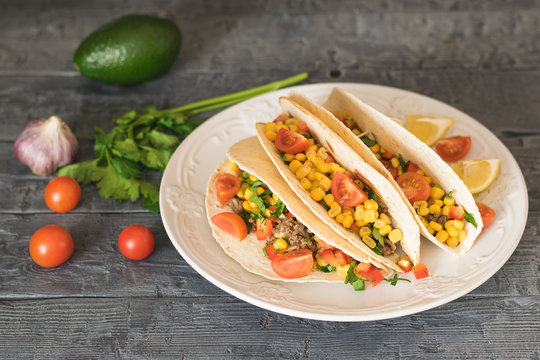 Freshly prepared Mexican tacos on a plate and avocado on a dark table with vegetables.