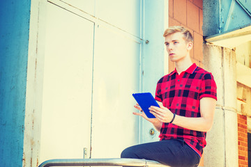 Young blonde American Man wearing red patterned Polo shirt, black pants, sitting on railing in front of office door way on street in New York, reading blue tablet computer, looking up, thinking..