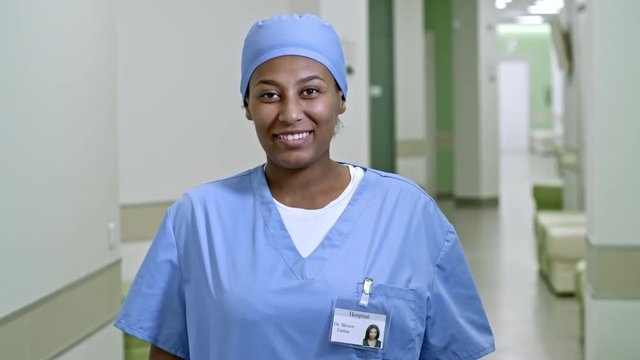 PAN of African female doctor in blue scrubs and cap standing in hospital hallway and smiling for camera; name Emma Brown is used in fictitious manner