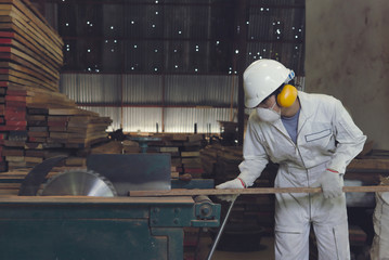 Vintage toned image of professional young Asian worker in white uniform and safety equipment cutting a piece of wood on table saw machine in carpentry factory.