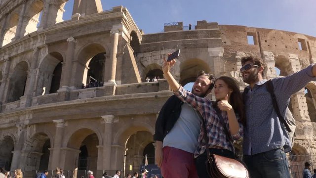 Three young friends tourists standing in front of colosseum in rome taking selfies with smartphone with backpacks sunglasses happy beautiful girl long hair slow motion
