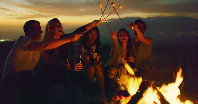 Group of friends relaxing around campfire roasting marshmallows, sunset bonfire lifestyle