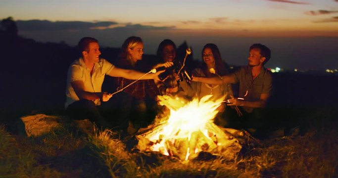 Group of friends relaxing around campfire roasting marshmallows, sunset bonfire lifestyle