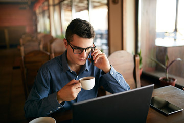 Caucasian businessman using phone while working on laptop and holding cup of coffee in hand. Multitasking concept. Busy freelancer in glasses drinks tea and talking on cellphone in cafe. Smart casual.