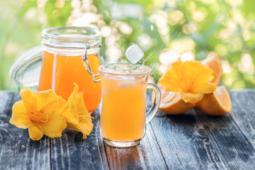 Freshly squeezed orange juice in a glass with slices of orange and yellow lilies flowers on the green bokeh background at Sunny .