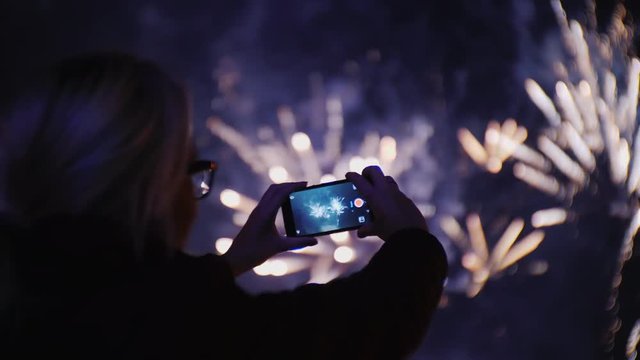 A woman takes pictures of fireworks in the night sky. Uses a smartphone