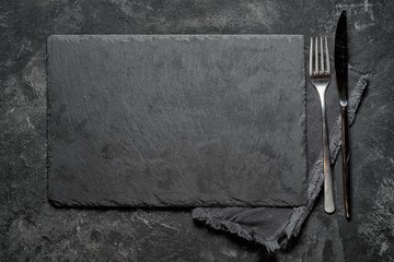 black stone board with fork and knife on black textured cement background, top view vith copy space...
