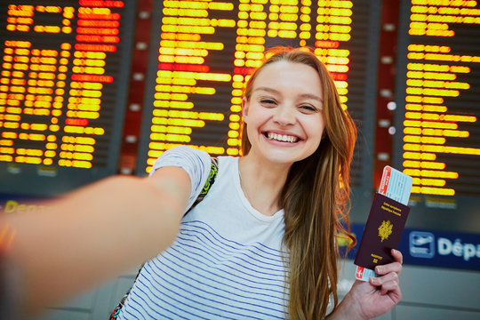 Girl in international airport, taking funny selfie with passport and boarding pass near flight information board