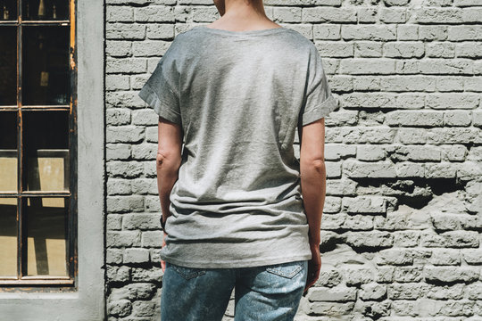 Summer day. Back view. Young millennial woman dressed in gray t-shirt is stands against gray brick wall. Mock up. Space for logo, text, image.