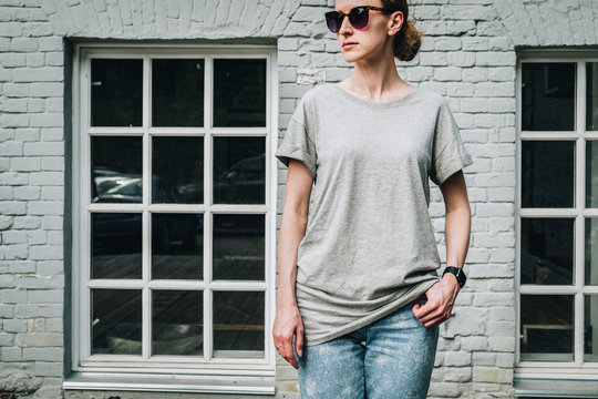 Summer day. Front view. Young millennial woman dressed in gray t-shirt is stands against gray brick wall. Mock up. Space for logo, text, image.