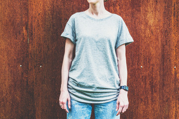 Summer day. Front view. Young millennial woman dressed in gray t-shirt is stands against wall of rusty metal. Mock up. Space for logo, text, image.