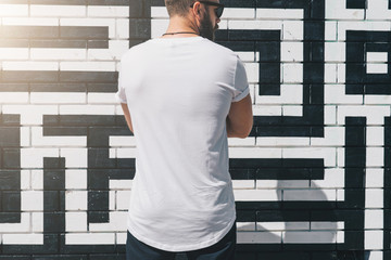 Summer day. Back view. Young bearded millennial man dressed in white t-shirt is stands against...