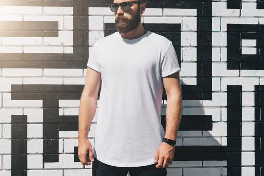 Summer day. Front view. Young bearded millennial man dressed in white t-shirt and sunglasses is stands against brick wall. Mock up. Space for logo, text, image. Instagram filter, film effect, bokeh
