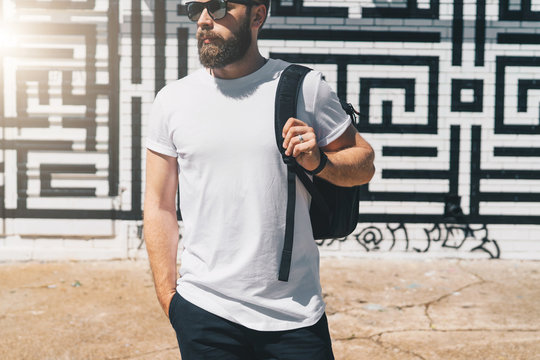 Summer day. Front view. Young bearded millennial man dressed in white t-shirt and sunglasses is stands against brick wall. Mock up. Space for logo, text, image. Instagram filter, film effect, bokeh