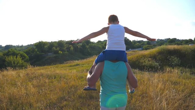 Father and son playing on summer field at sunny day. Little boy sitting on shoulders of his daddy and playing raised hands as airplane. Parent with kid having fun together. Slow motion Rear back view