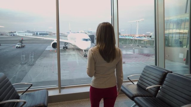 A young, beautiful girl approaches the panoramic window of the airport through which the aircraft is seen