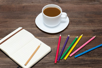 Coffee mug, colored pencils and a notebook. Wooden background