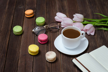 Fototapeta na wymiar Cup of tea, macarons, glasses, pink tulips and notebook on wooden background.