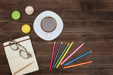 Top viev on a coffee mug, macaroons, colored pencils and a notebook. Wooden background