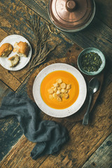 Flat-lay of fall warming pumpkin cream soup with croutons and seeds on board over rustic wooden background, top view, vertical composition. Autumn vegetarian, vegan, healthy comfort food concept