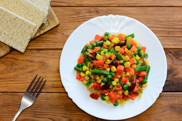 Healthy vegetable stew on a white plate and a wooden background. Stewed vegetables mix. Stewed carrots, green beans, red beans, peas, corn, pepper onions. Top view