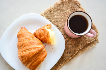 Close up of breakfast, cup of coffee and big tasty croissant on white plate on white table.