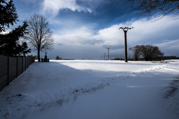 a snowy landscape with a cross under a tree and a pole with an electric lead in winter