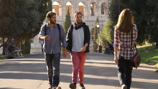 Young lovely gay couple tourists walk in park road with trees colosseum in background in rome at sunset holding hands. Boyfriend gets jealous when beautiful girl walks by glancing. Slow motion.