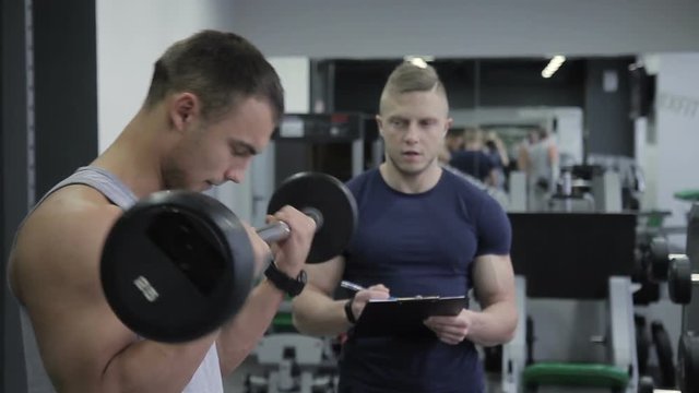Sportsman is doing barbell biceps curl and his male trainer is controlling his effort. Athlete is standing in the gym and doing exercise for his muscules by the weight equipment in his hands
